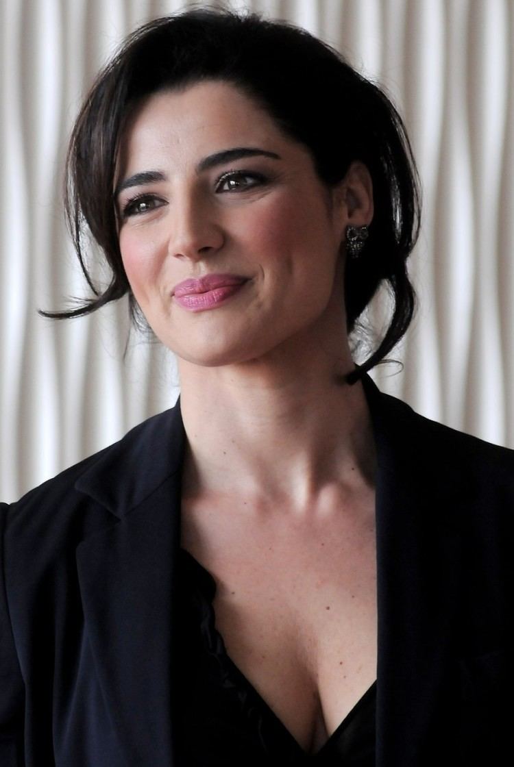 Luisa Ranieri with a tight-lipped smile while looking afar with a black messy hair and wearing earrings and a black blouse under a black coat that exposes her cleavage