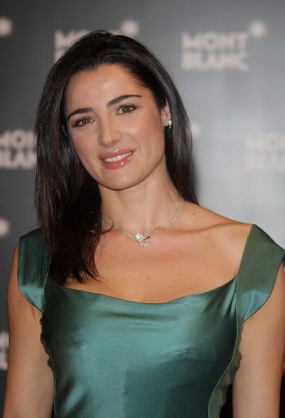 Luisa Ranieri smiling with black shoulder-length hair while wearing a green sleeveless blouse, earrings, and necklace