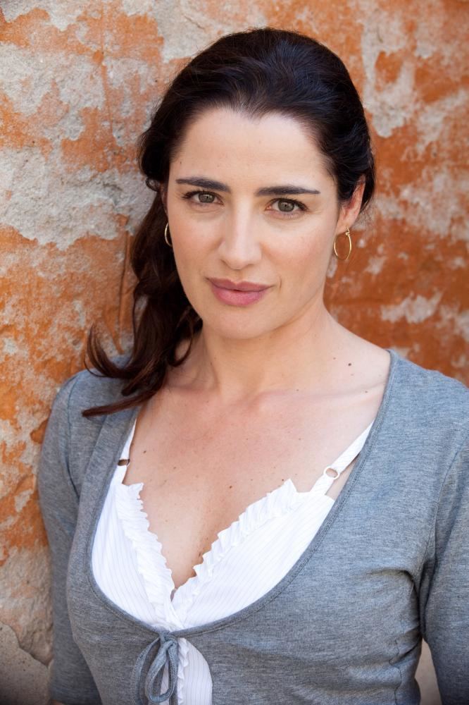 Luisa Ranieri with a tight-lipped smile and tied-up hair while wearing earrings and a white blouse with a sweetheart neckline under a gray blazer