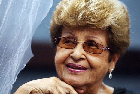 Lydia Echevarría smiling while hand on her chin and wearing a black blouse, sunglasses, and earrings