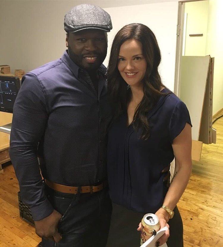 Lydia Hull smiling while holding a drink and wearing a navy blue blouse together with 50 Cent in a blue long sleeve and gray cap