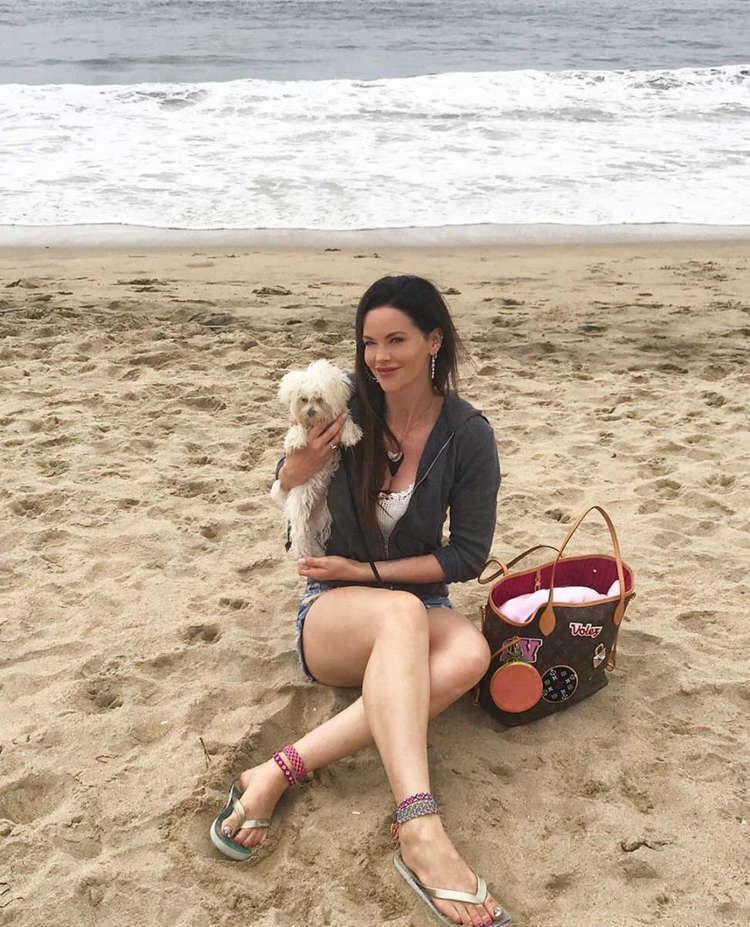 Lydia Hull smiling, sitting on the beach and carrying a dog while wearing a white blouse under a gray hoodie jacket and denim shorts