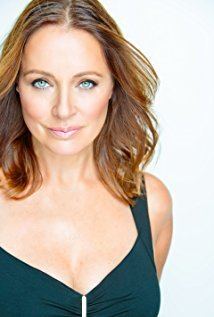 Lydie Denier with a tight-lipped smile and golden brown wavy hair while wearing a blue-green sleeveless blouse