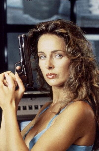 Lydie Denier holding a gun with a fierce look and curly hair while wearing a blue brassiere