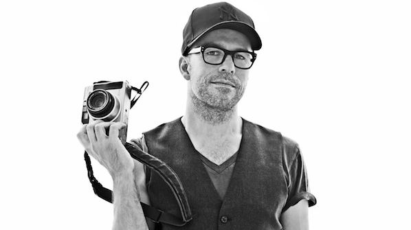Lyle Owerko with a serious face while holding a camera, wearing eyeglasses, a cap, a vest, and a shirt.