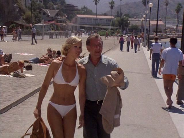 Lynette Mettey with short blonde hair, wearing a white swimsuit and carrying a brown bag with a man wearing long sleeves and pants holding his suit at the beach.