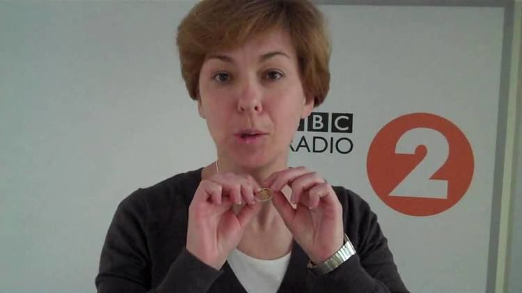 Lynn Bowles is talking while holding her necklace, with short blonde hair, wearing a watch, and a black blazer over a white shirt.