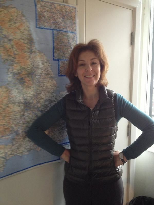 Lynn Bowles smiling while her hands are on her waist, with shoulder-length wavy hair, wearing a watch, a black vest over a green long sleeve shirt, and black pants.