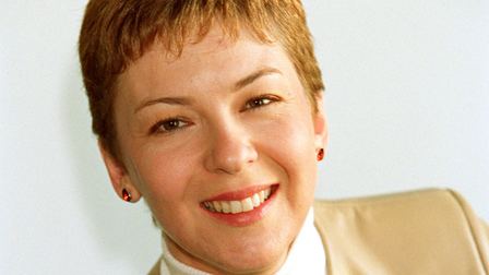 Lynn Bowles smiling, with short blonde hair, wearing earrings and a brown coat over a white top.