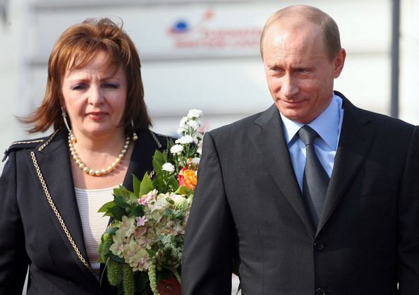 Lyudmila Putina and Vladimir Putin with a tight-lipped smile while Lyudmila is holding a bouquet of flowers and wearing pearl necklace, earrings & a black coat with white inner blouse