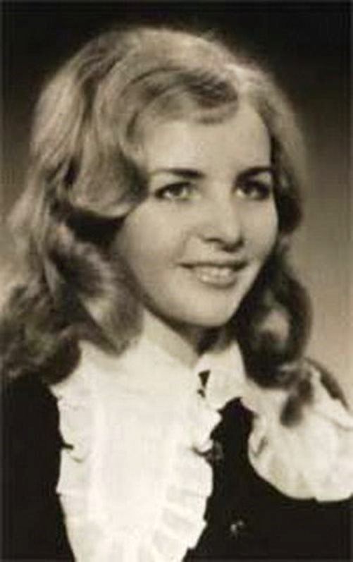Lyudmila Putina smiling while looking at something, with blonde wavy hair, and wearing a black and white blouse
