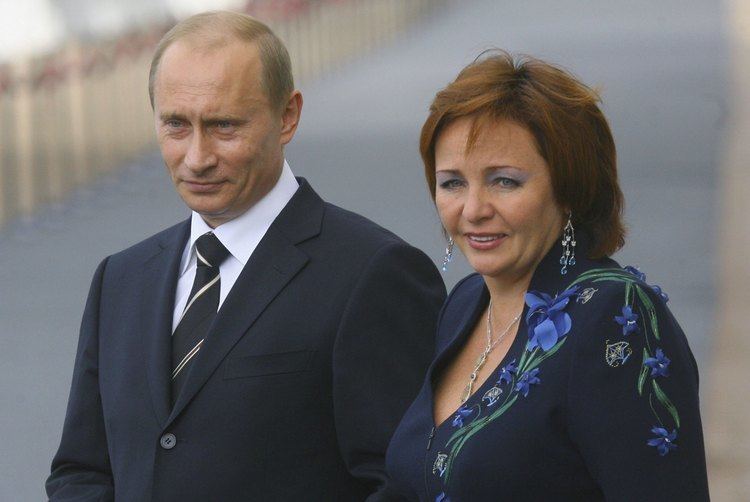 Vladimir Putin and Lyudmila Putina are smiling and looking afar. Vladimir is wearing a dark blue coat, white long sleeve, and black striped necktie while Lyudmila is wearing a blue floral blouse