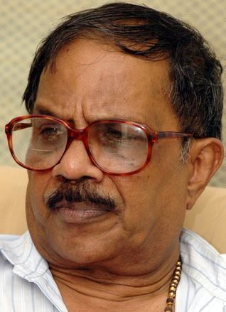 M. T. Vasudevan Nair looking at something and wearing a striped polo shirt along with a bead necklace and red eyeglasses.