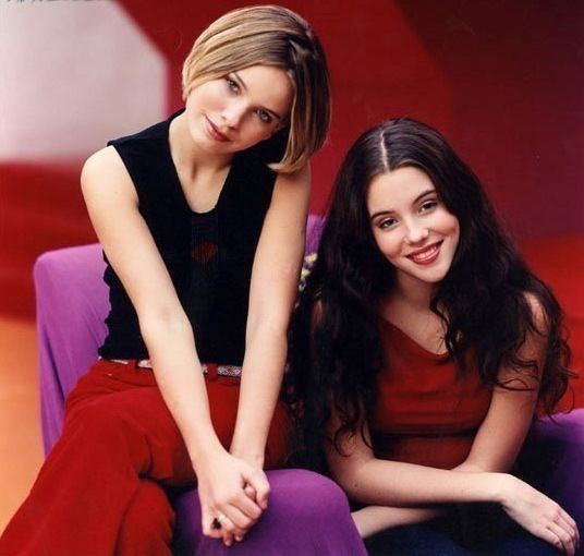 Marit Larsen and Marion Raven of the M2M band are both smiling and sitting on a violet chair while Marit Larsen is wearing a black top and Marion is wearing a red top