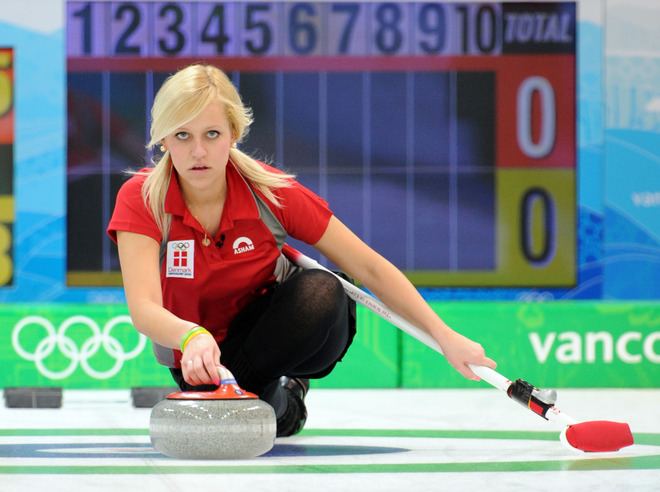 Madeleine Dupont looking afar and holding a polished granite stone and broom while wearing a red polo shirt and black pants