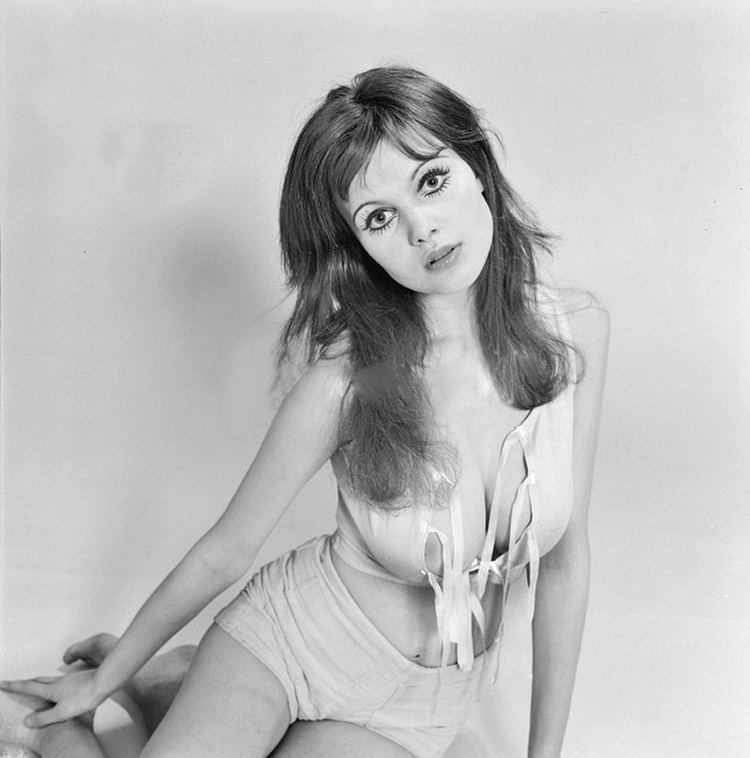 Madeline Smith sitting on the floor while wearing a shorts and sleeveless top showing off her cleavage