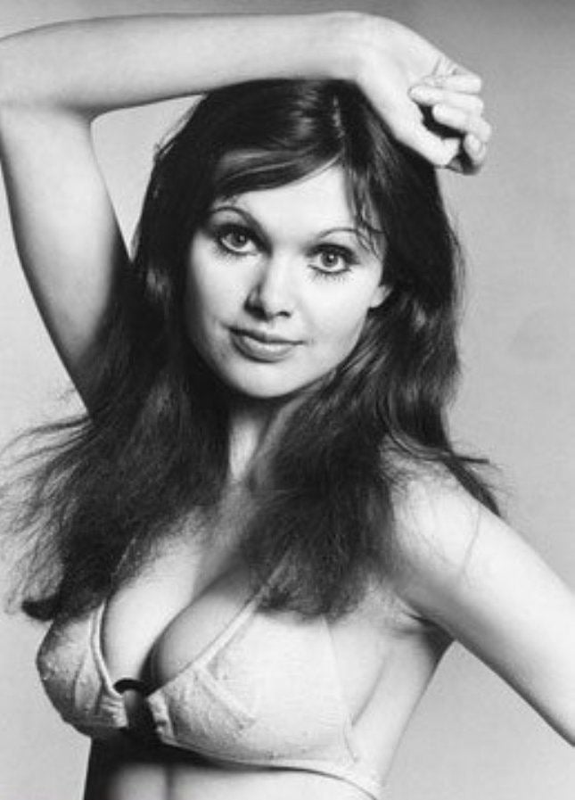 Madeline Smith with a tight-lipped smile while her hand is on her head and she is wearing a brassiere
