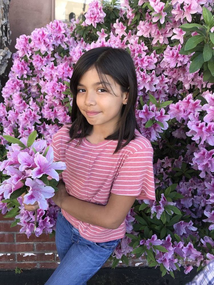 Madelyn Miranda with a cute smile while holding and surrounded with flowers, with short hair, wearing a striped pink shirt and blue jeans.