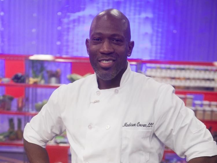 Madison Cowan smiling with a mustache, beard, stud earrings, bald head, a shelf in the background, and arms on his waist while wearing Chef's uniform the white double-breasted jacket with his name on the right side