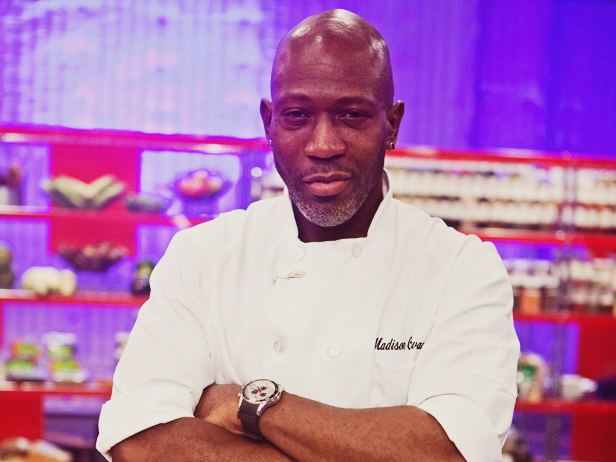 Madison Cowan with a tight-lipped smile, mustache, beard, stud earrings, bald head, a shelf in the background, and his arms crossed while wearing a wristwatch and Chef's uniform the white double-breasted jacket with his name on the right side