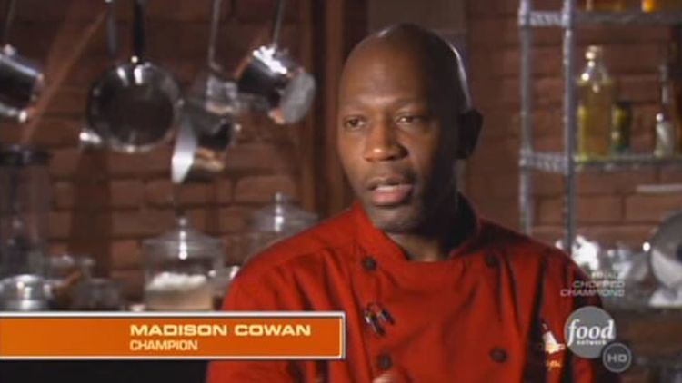 Madison Cowan talking to the interviewer with a serious face and cooking materials behind him while he is wearing a red double-breasted jacket