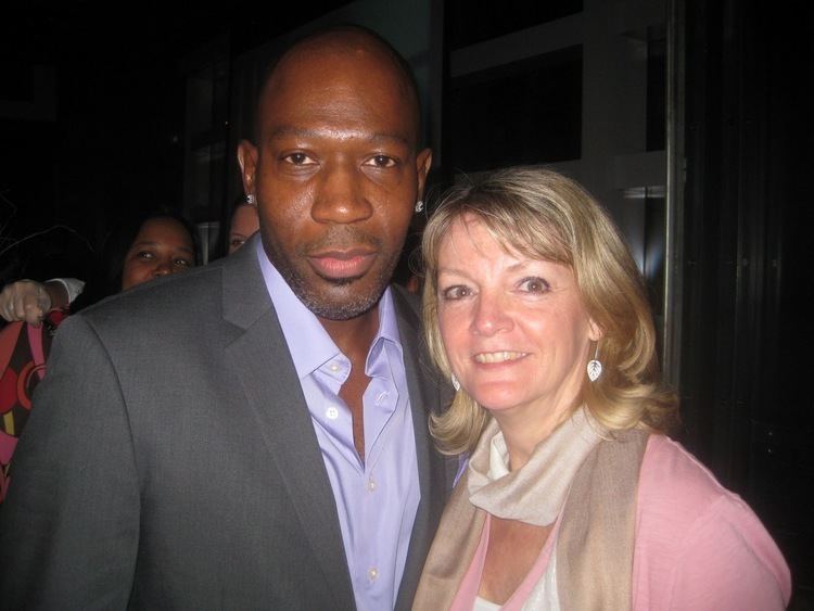 Madison Cowan and the woman beside him are smiling. Madison with a mustache, beard, stud earrings, and bald head is wearing earrings and light purple long sleeves under a dark gray coat while the woman beside him is wearing earrings, a beige scarf, and a pink blouse