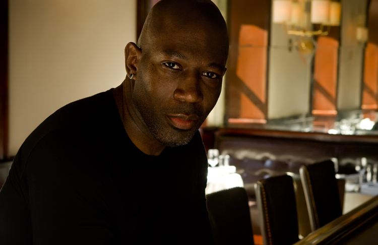 Madison Cowan with a tight-lipped smile, beard, bald head, and bar counter with chairs in the background. Madison is wearing earrings and a black t-shirt