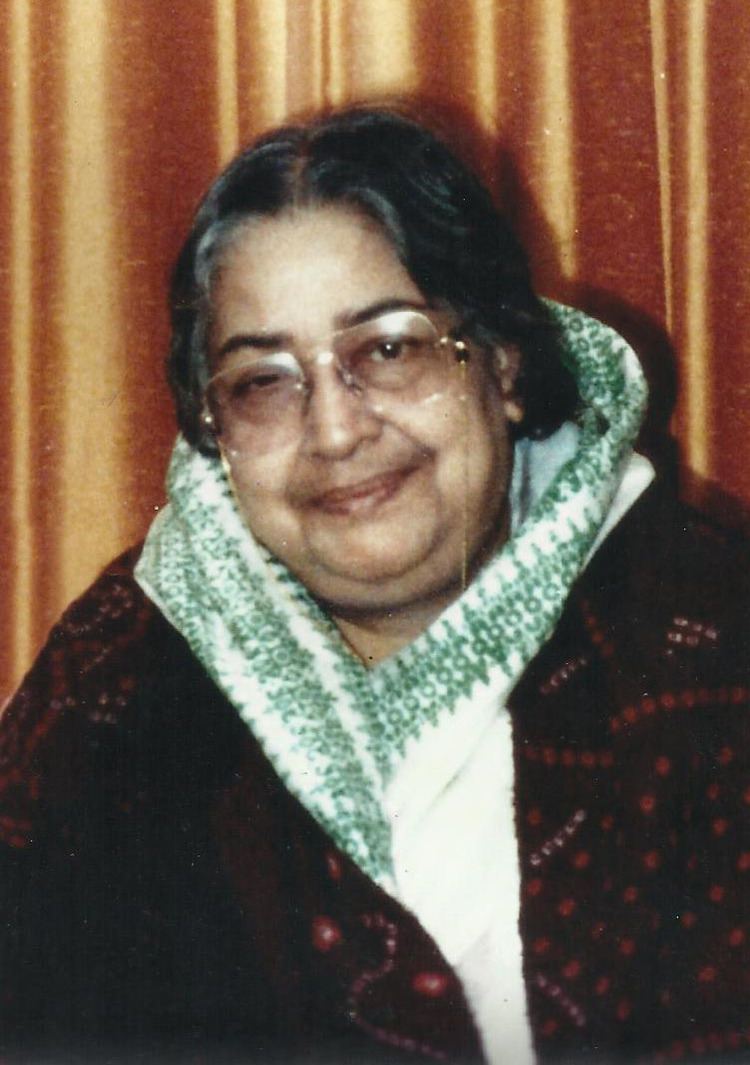 Maitreyi Devi with a tight-lipped smile while wearing a white and green scarf, maroon blazer, and eyeglasses
