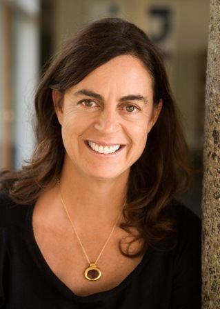 Maja Hoffmann smiling while leaning on the wall, with wavy hair, wearing a necklace, and a black shirt.