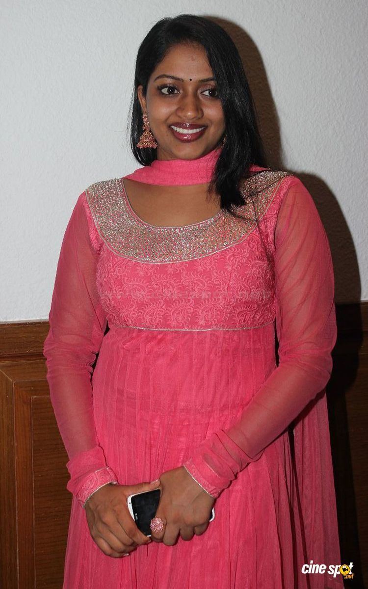 Mallika wearing earrings, a pink scarf, and a pink dress and holding a mobile phone.