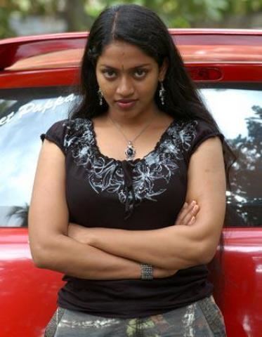 Mallika wearing earrings, a necklace, and a brown shirt beside a red car.