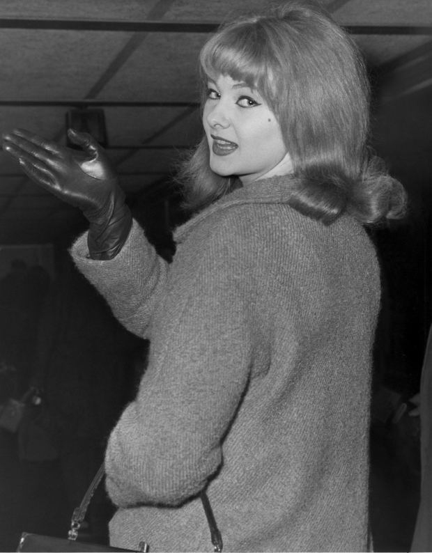 Mandy Rice-Davies posing while pointing at something and wearing a gray coat and black gloves and carrying a black bag.