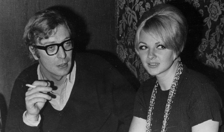 Mandy Rice-Davies sitting down with Michael Caine and wearing a black shirt and a necklace.