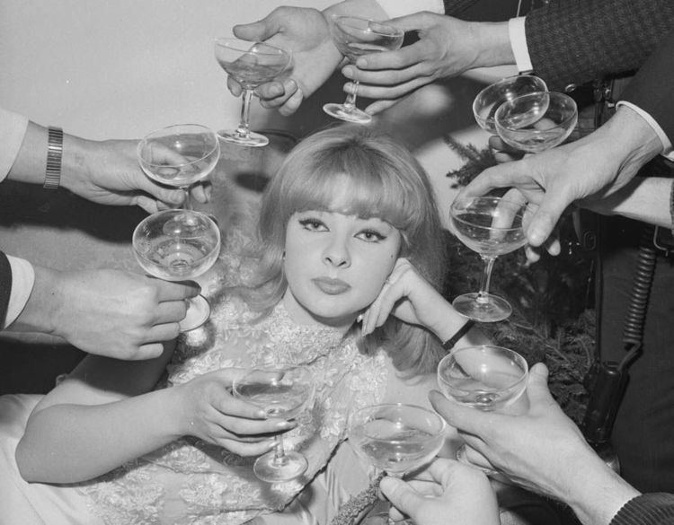 Mandy Rice-Davies posing with her hand in her cheek and surrounded by men handing her glasses.