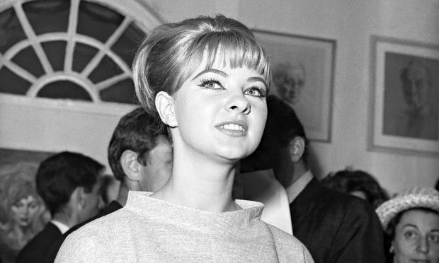 Mandy Rice-Davies smiling at a gathering with bangs in her hair and wearing a light gray dress.