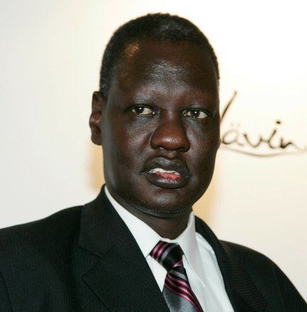 Manute Bol looking afar while wearing a black coat, white long sleeves, and gray and pink striped necktie