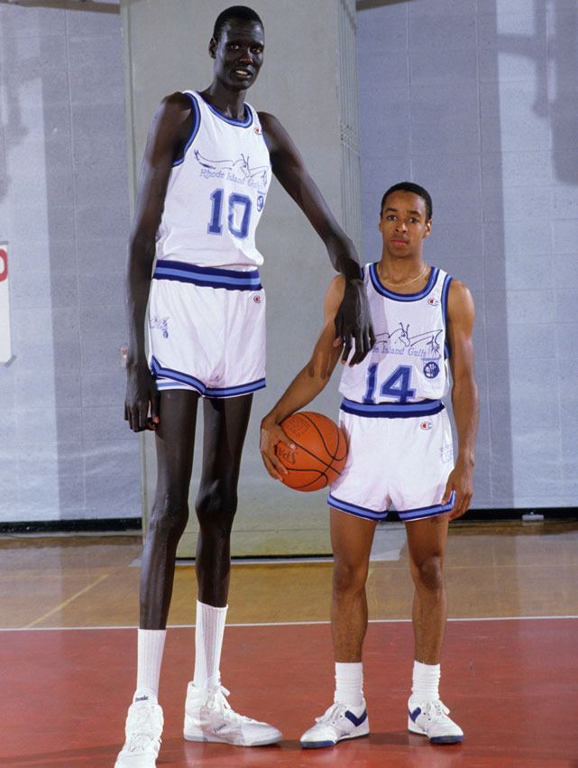 Manute Bol smiling and leaning on Spud Webb's shoulder while wearing a white and blue jersey with the number 10 and white rubber shoes