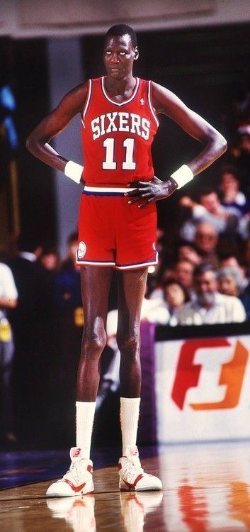 Manute Bol's hand on his hips while wearing the white and red Sixers jersey with the number 11 and white and red rubber shoes