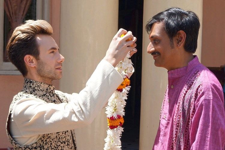 Manvendra Singh Gohil smiling and wearing purple long sleeves with a man with a serious face, blonde hair, and holding a floral necklace.