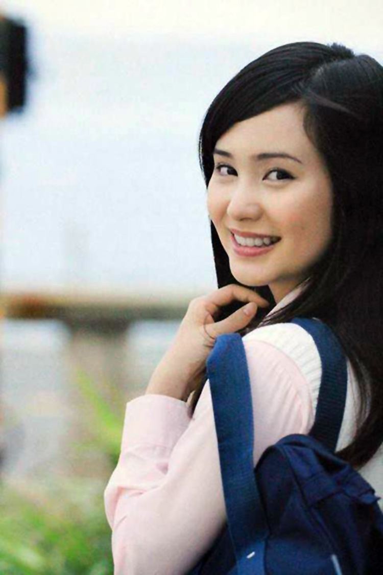 Mao Ichimichi smiling while carrying a blue bag and wearing a light pink long sleeve blouse and white vest