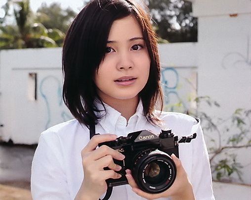 Mao Ichimichi with a tight-lipped smile while holding a DSLR camera and wearing a white blouse