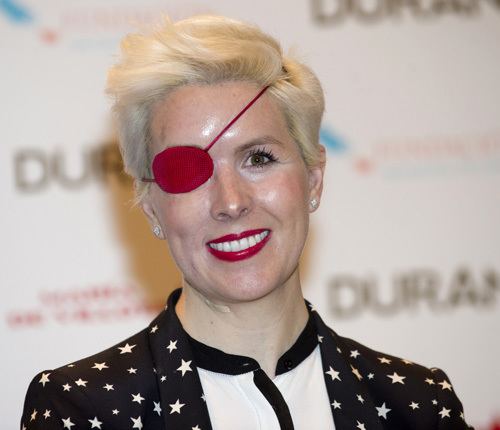 María de Villota smiling, with blonde short hair, and wearing a red eye patch, a black and white coat with stars design, and a black and white blouse