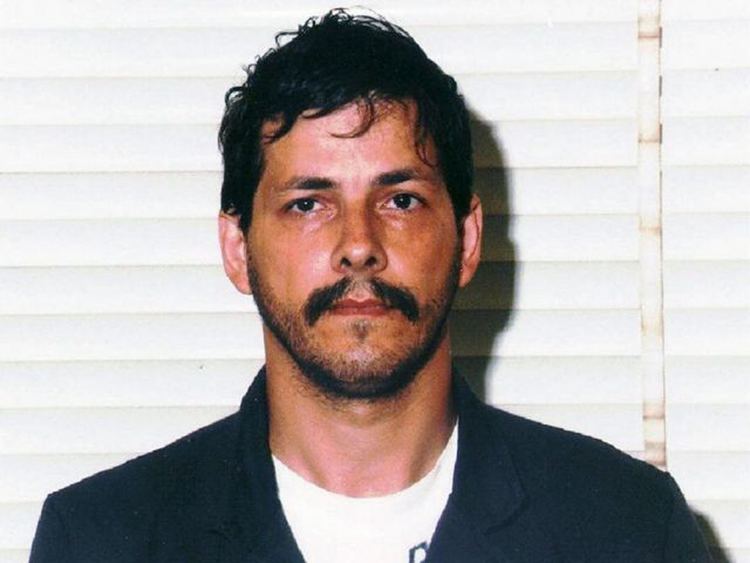 Marc Dutroux is serious, in front of a white blinds, has black hair, mustache and beard, wearing white top under a black shirt.