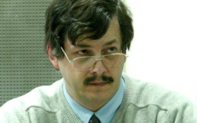 Marc Dutroux is serious, sitting staring to his left, has black hair, mustache wearing eyeglasses, blue polo with tie, and gray long sleeve.