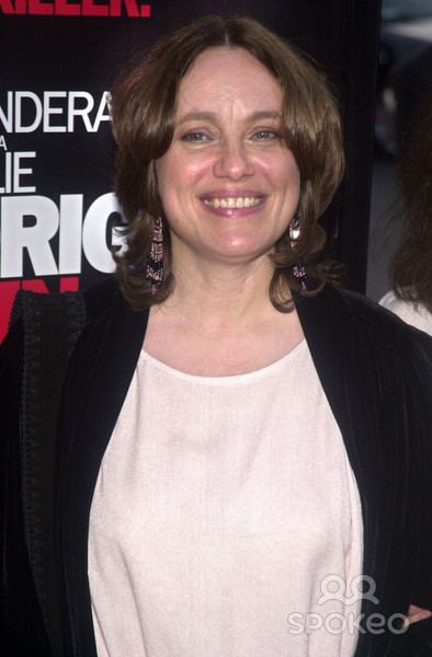 Marcheline Bertrand smiling with wavy hair and wearing a white blouse under a black cardigan and dangling earrings