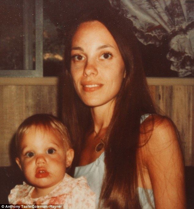Marcheline Bertrand smiling with her long hair down and carrying a baby and wearing a white sleeveless shirt and a necklace