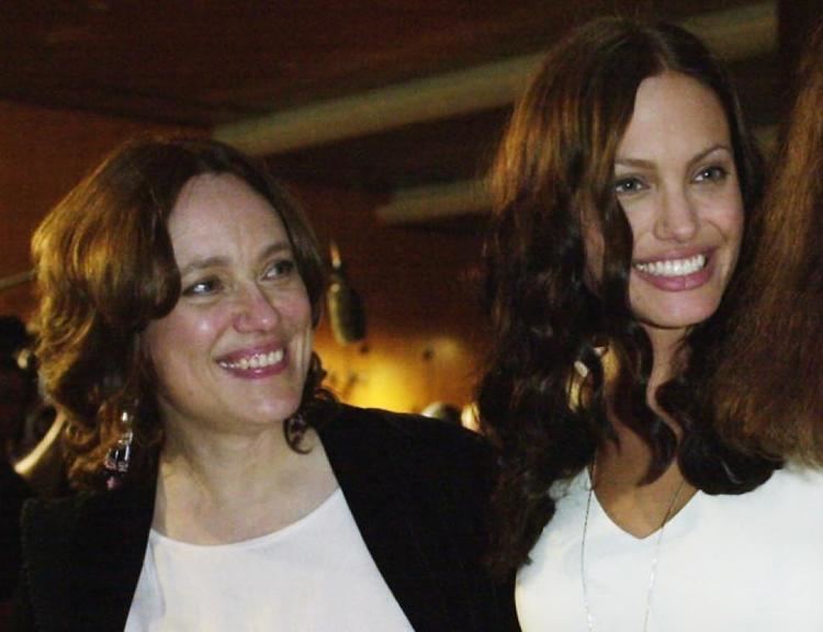 Marcheline Bertrand smiling with wavy hair and wearing a white blouse under a black cardigan and dangling earrings along with Angeline Jolie having curly hair and wearing a white blouse and necklace
