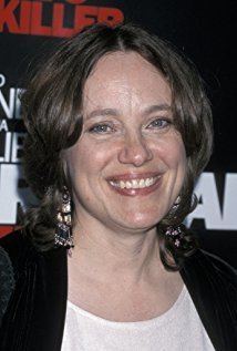 Marcheline Bertrand smiling with wavy hair and wearing a white blouse under a black cardigan and dangling earrings
