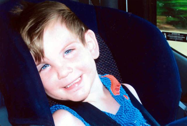 Marcus Fiesel is smiling, sitting on a car seat, has brown hair, blue eyes, wearing a blue shirt.
