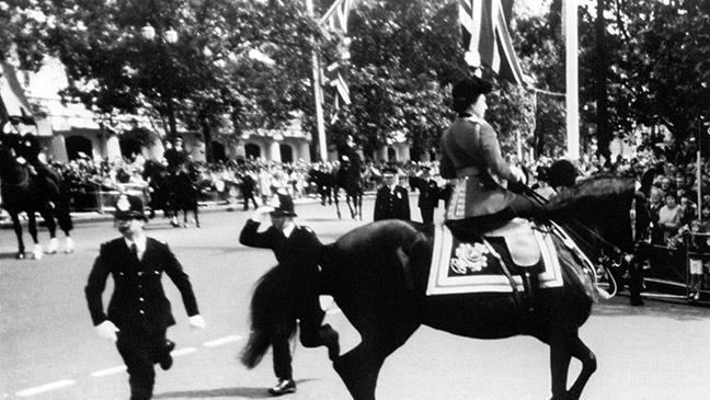 Queen Elizabeth II on horseback after shots were heard as she rode down the Mall in 1981, along with other troops running in the background wearing red tunics and bearskin hats. Queen Elizabeth is wearing red tunics and bearskin hats
